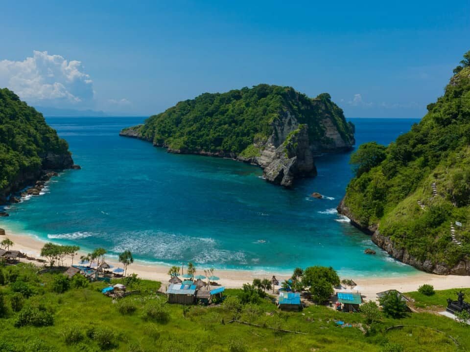Tropical cove with turquoise water, white sand beach, and lush green cliffs; small beach huts line the shore.