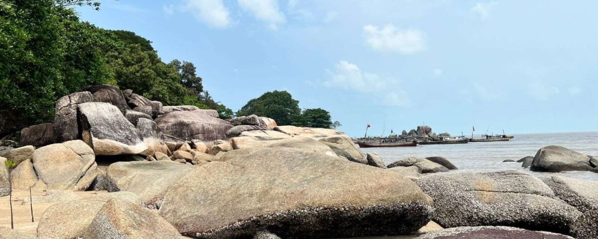 Rocky beach with large weathered boulders, trees on the left, and calm sea with distant boats offshore.