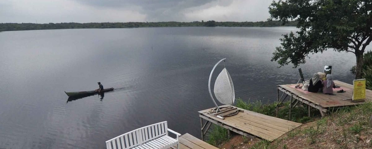 Person paddling a small canoe on a calm, overcast lake with a distant shoreline visible under a cloudy sky.