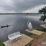 Person paddling a small canoe on a calm, overcast lake with a distant shoreline visible under a cloudy sky.