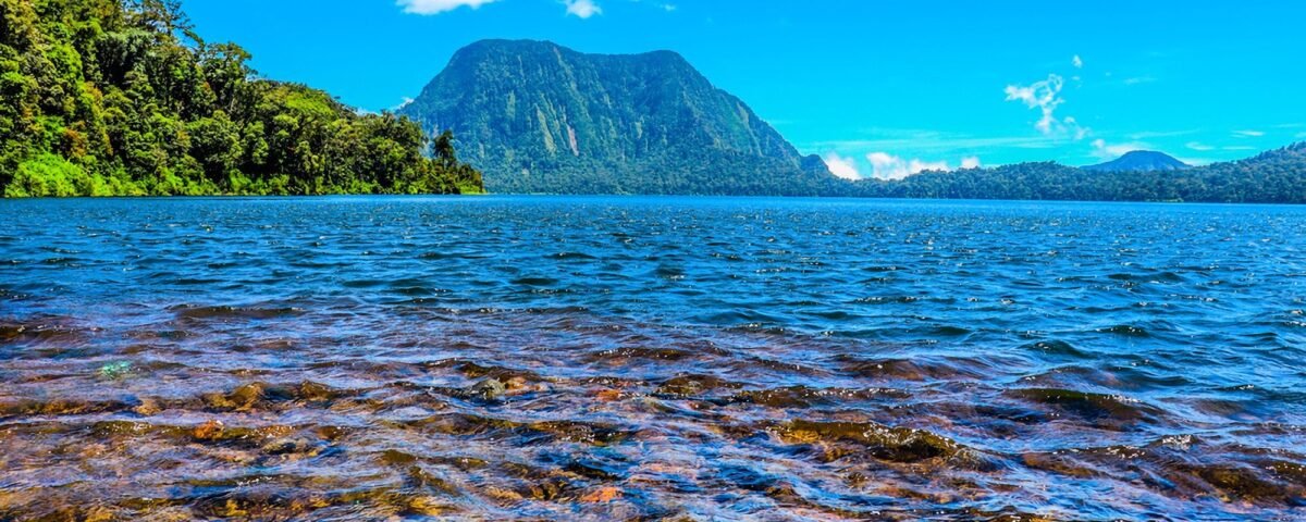 Tropical lake with clear blue water, rocky foreground, forested left shore, and a tall mountain under a bright sky.