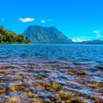 Tropical lake with clear blue water, rocky foreground, forested left shore, and a tall mountain under a bright sky.