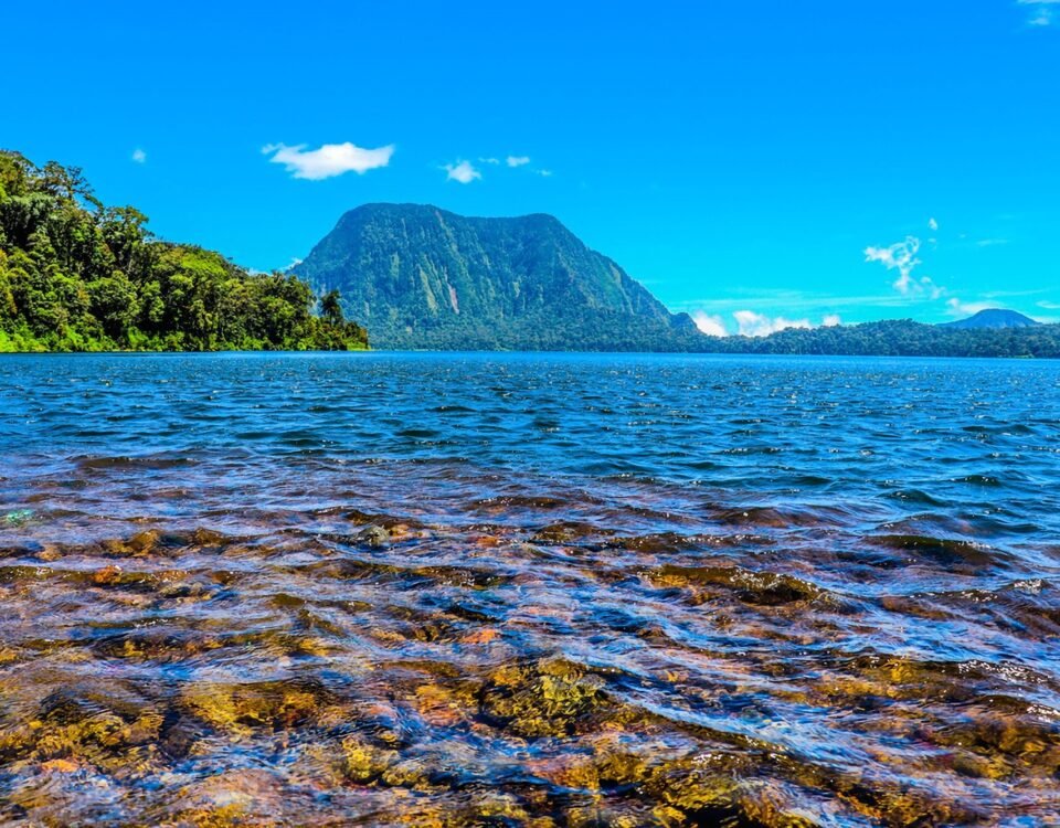 Tropical lake with clear blue water, rocky foreground, forested left shore, and a tall mountain under a bright sky.