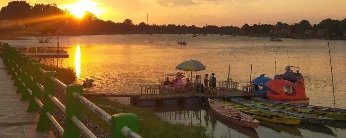 Sunset over a calm river with a green railing, a pier, and colorful pedal boats nearby as a group gathers on a floating platform under a green umbrella.