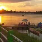 Sunset over a calm river with a green railing, a pier, and colorful pedal boats nearby as a group gathers on a floating platform under a green umbrella.