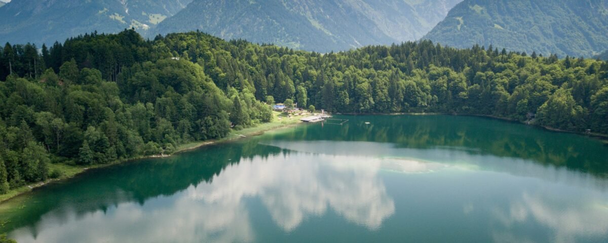 Alpine lake surrounded by dense green forest and towering mountains under a partly cloudy blue sky, with the clouds reflected on the water surface.