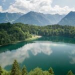 Alpine lake surrounded by dense green forest and towering mountains under a partly cloudy blue sky, with the clouds reflected on the water surface.