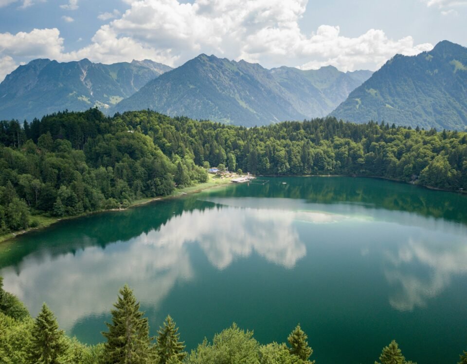Alpine lake surrounded by dense green forest and towering mountains under a partly cloudy blue sky, with the clouds reflected on the water surface.
