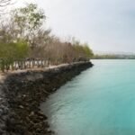 Path along a rocky breakwater beside turquoise water, with leafless trees and small shelter structures along the shore.”, “Seaside promenade with a stone jetty and bright blue-green water, trees lining the coast.”, “Coastal scene: rocky barrier, clear turquoise sea, and row of simple huts under trees.”, “Stone shoreline with calm turquoise water and a line of shade structures along the path.”