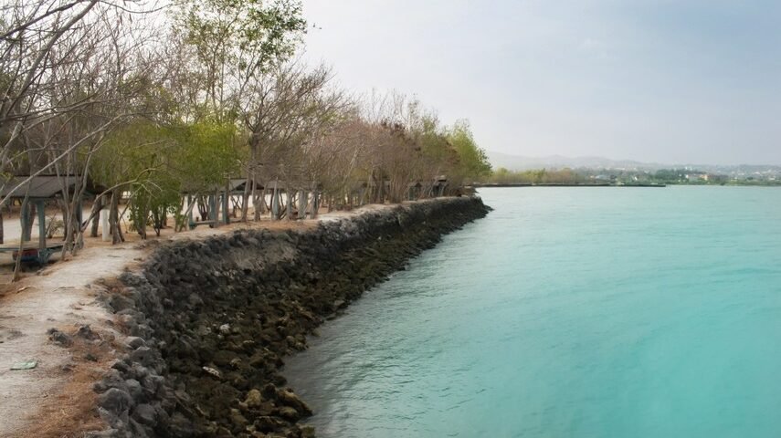 Path along a rocky breakwater beside turquoise water, with leafless trees and small shelter structures along the shore.”, “Seaside promenade with a stone jetty and bright blue-green water, trees lining the coast.”, “Coastal scene: rocky barrier, clear turquoise sea, and row of simple huts under trees.”, “Stone shoreline with calm turquoise water and a line of shade structures along the path.”