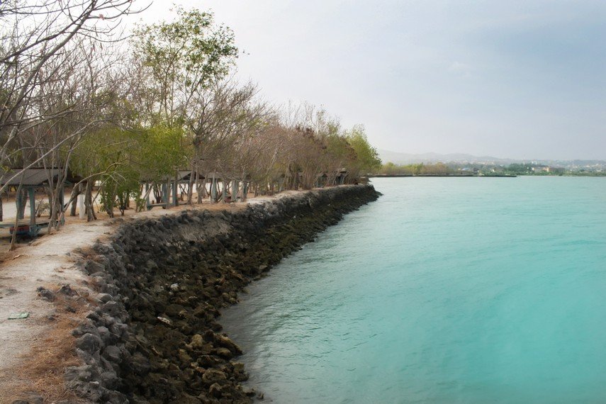 Path along a rocky breakwater beside turquoise water, with leafless trees and small shelter structures along the shore.”, “Seaside promenade with a stone jetty and bright blue-green water, trees lining the coast.”, “Coastal scene: rocky barrier, clear turquoise sea, and row of simple huts under trees.”, “Stone shoreline with calm turquoise water and a line of shade structures along the path.”