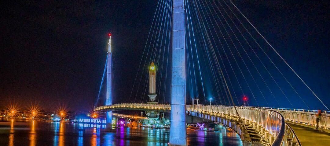 Night view of a lit cable-stayed bridge over a calm river, blue and white lights reflecting on the water at night.