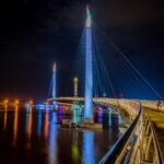 Night view of a lit cable-stayed bridge over a calm river, blue and white lights reflecting on the water at night.