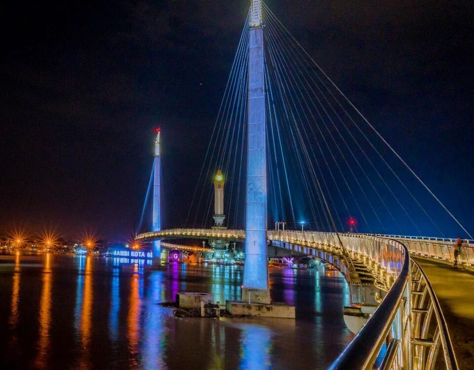 Night view of a lit cable-stayed bridge over a calm river, blue and white lights reflecting on the water at night.