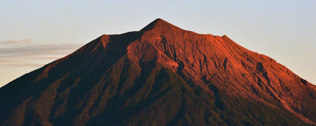 Sunlit mountain peak with red-orange slopes and dark textured ridges against a pale blue sky.