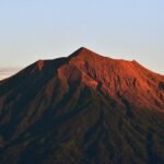 Sunlit mountain peak with red-orange slopes and dark textured ridges against a pale blue sky.