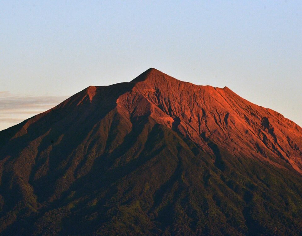 Sunlit mountain peak with red-orange slopes and dark textured ridges against a pale blue sky.