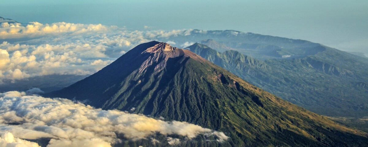 Snow-dusted volcanic peak rising above a sea of fluffy white clouds with distant mountain ridges in the background