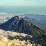 Snow-dusted volcanic peak rising above a sea of fluffy white clouds with distant mountain ridges in the background