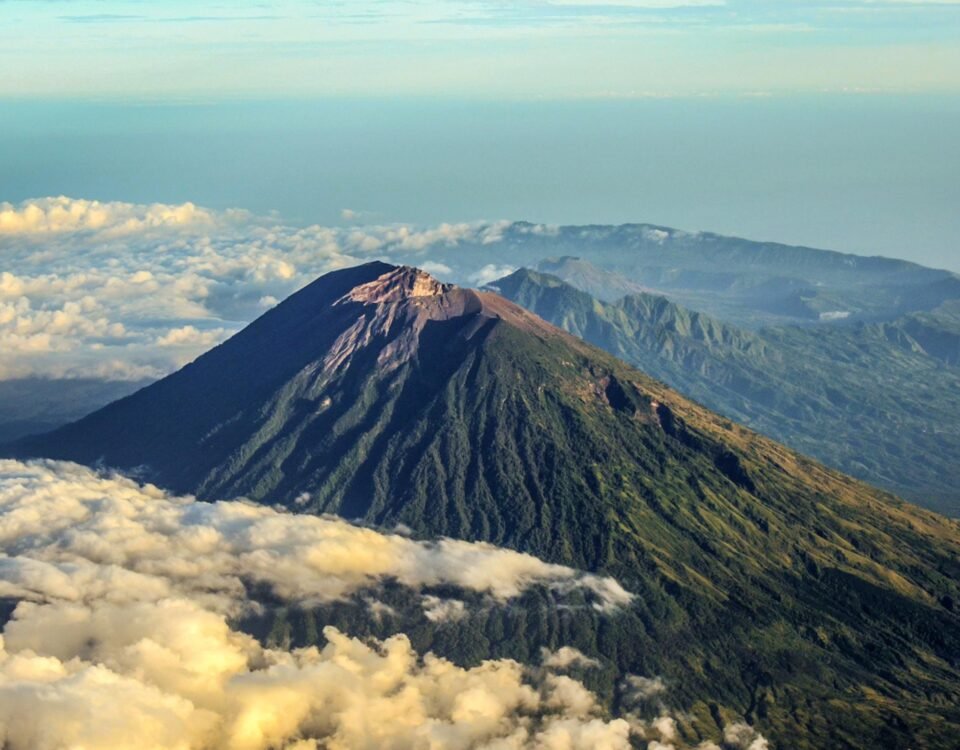 Snow-dusted volcanic peak rising above a sea of fluffy white clouds with distant mountain ridges in the background