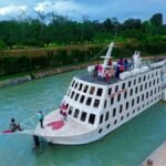 A white multi‑deck passenger ship with many windows sits in a calm green canal, passengers on the upper deck and a gangway connecting to shore nearby.