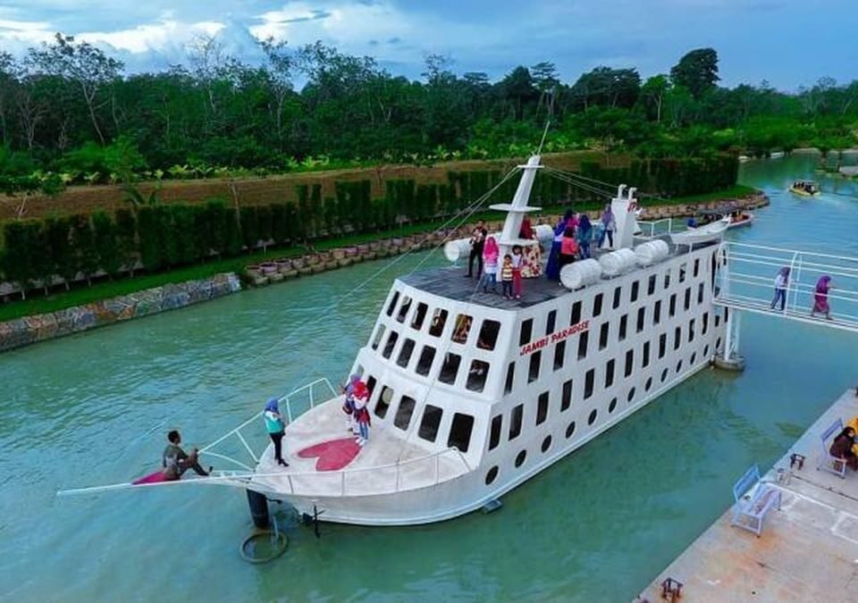 A white multi‑deck passenger ship with many windows sits in a calm green canal, passengers on the upper deck and a gangway connecting to shore nearby.