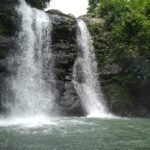 Two-tier waterfall cascading over a rocky ledge into a greenish pool, surrounded by dense jungle foliage.