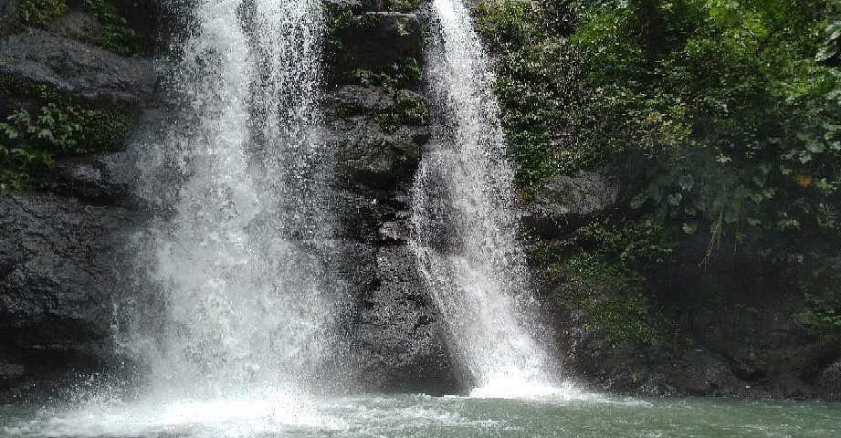 Two-tier waterfall cascading over a rocky ledge into a greenish pool, surrounded by dense jungle foliage.