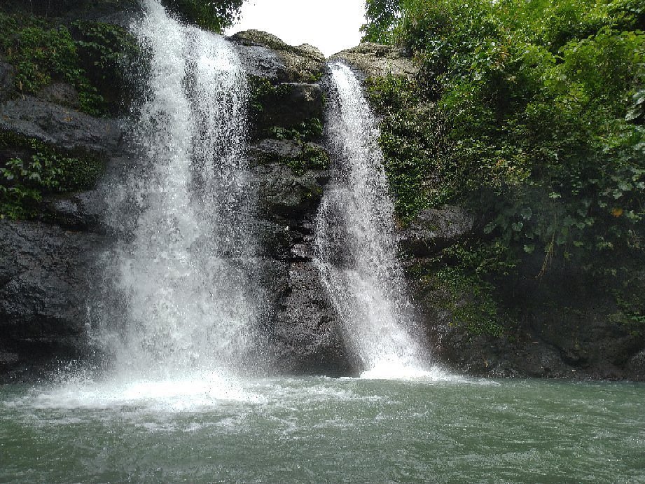 Two-tier waterfall cascading over a rocky ledge into a greenish pool, surrounded by dense jungle foliage.