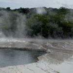 Geothermal hot spring with steaming pool and white travertine terraces; a forested hillside in the background.