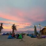 Beach scene at sunset with string lights, palm trees, and people relaxing on colorful bean bags near a thatched hut.