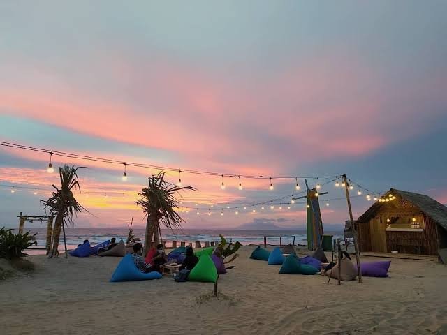 Beach scene at sunset with string lights, palm trees, and people relaxing on colorful bean bags near a thatched hut.