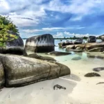 Tropical beach with large granite boulders, white sand, and turquoise water under a blue sky.