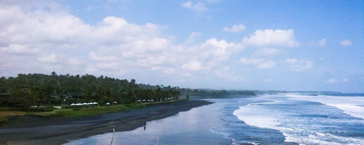 A tropical beach with dark sand, palm trees along the shore, and rolling waves under a partly cloudy sky.