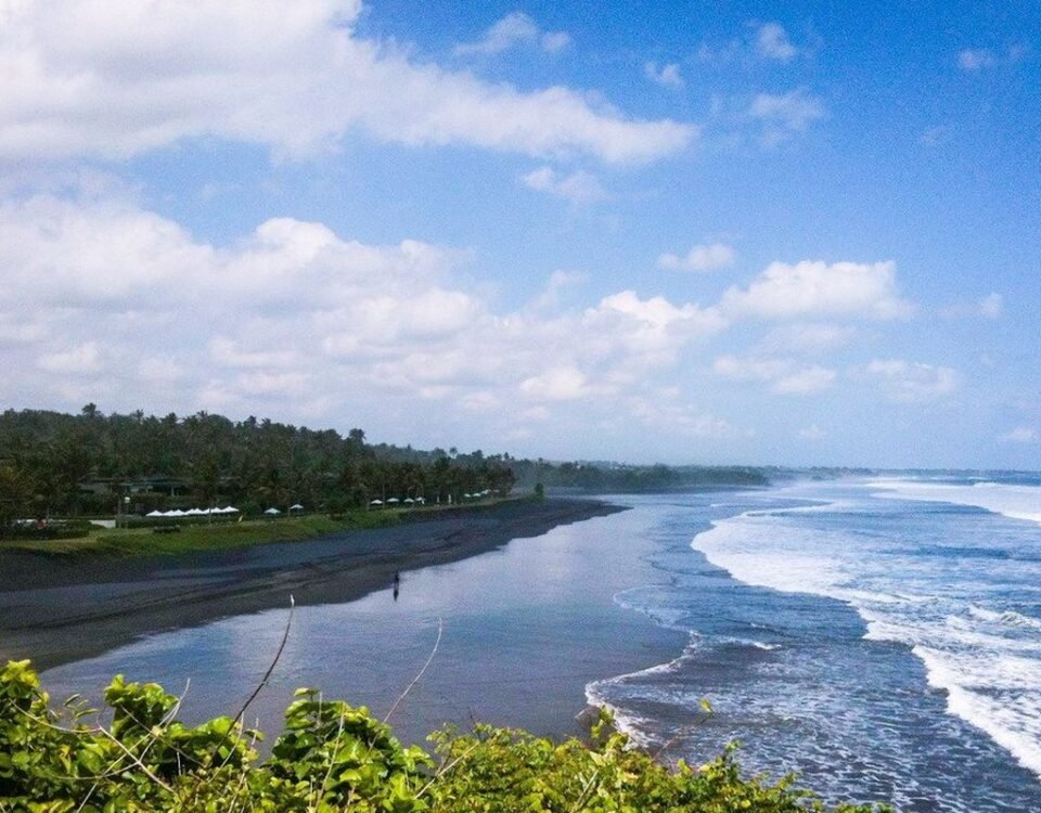 A tropical beach with dark sand, palm trees along the shore, and rolling waves under a partly cloudy sky.