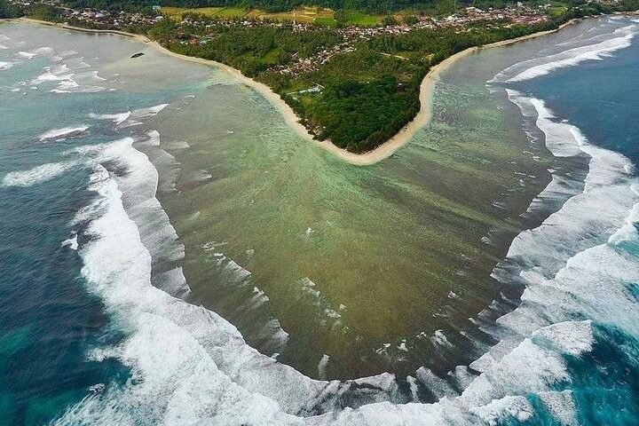 Aerial view of a coastal heart-shaped inlet formed by a sandy beach and green vegetation, with turquoise water and white waves along the shore.