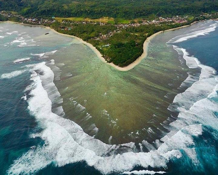 Aerial view of a coastal heart-shaped inlet formed by a sandy beach and green vegetation, with turquoise water and white waves along the shore.