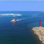 Red lighthouse atop a rocky breakwater extending into calm blue sea, with a small red boat nearby.
