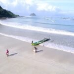 Sandy beach with a small green outrigger canoe at the water’s edge; two children play near the shore, hills on the left and distant boats on the calm sea.