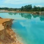 Turquoise lake edged by orange-brown rocky cliffs, with trees in the distance under a blue sky.