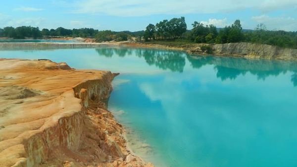 Turquoise lake edged by orange-brown rocky cliffs, with trees in the distance under a blue sky.