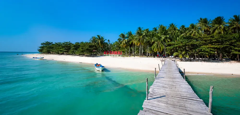 Tropical beach with turquoise water, white sand, and a wooden pier leading to palm trees along the shore.