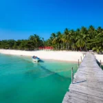 Tropical beach with turquoise water, white sand, and a wooden pier leading to palm trees along the shore.