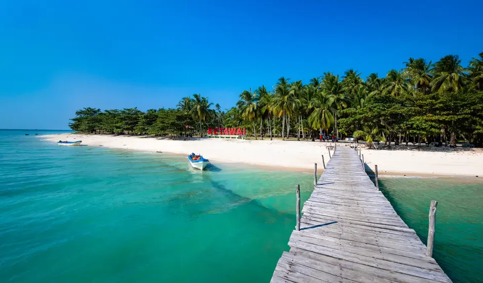 Tropical beach with turquoise water, white sand, and a wooden pier leading to palm trees along the shore.