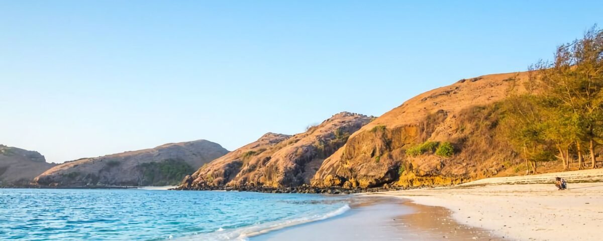 Wide coastal scene with clear blue sky, rocky orange cliffs, and a calm turquoise sea at the shore; a person sits on the sand.