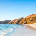 Wide coastal scene with clear blue sky, rocky orange cliffs, and a calm turquoise sea at the shore; a person sits on the sand.