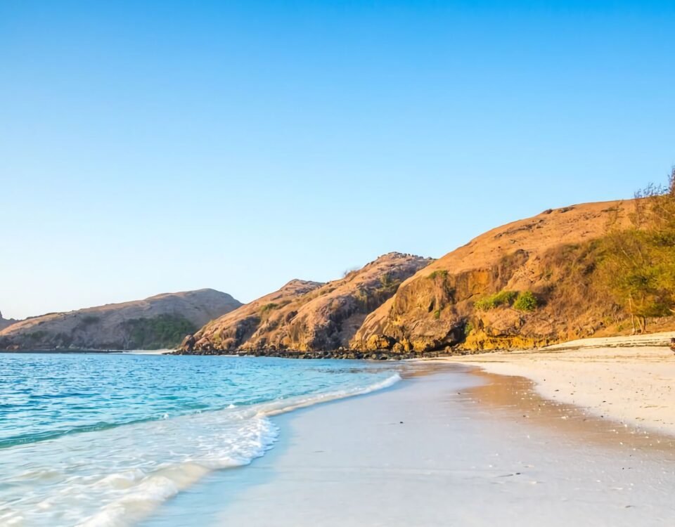 Wide coastal scene with clear blue sky, rocky orange cliffs, and a calm turquoise sea at the shore; a person sits on the sand.