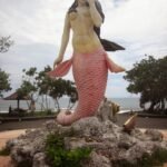 Life-size mermaid statue with a pink scaled tail on a rocky pedestal by the seaside park backdrop.