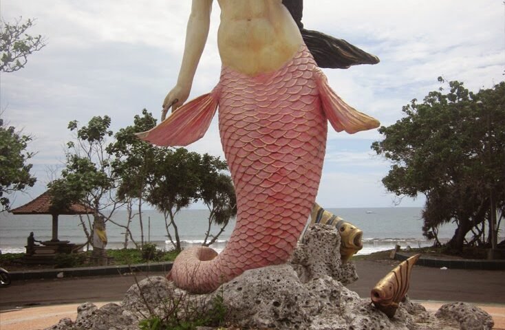 Life-size mermaid statue with a pink scaled tail on a rocky pedestal by the seaside park backdrop.