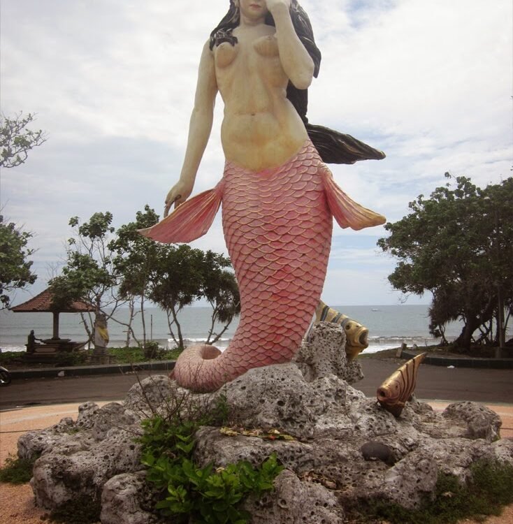Life-size mermaid statue with a pink scaled tail on a rocky pedestal by the seaside park backdrop.